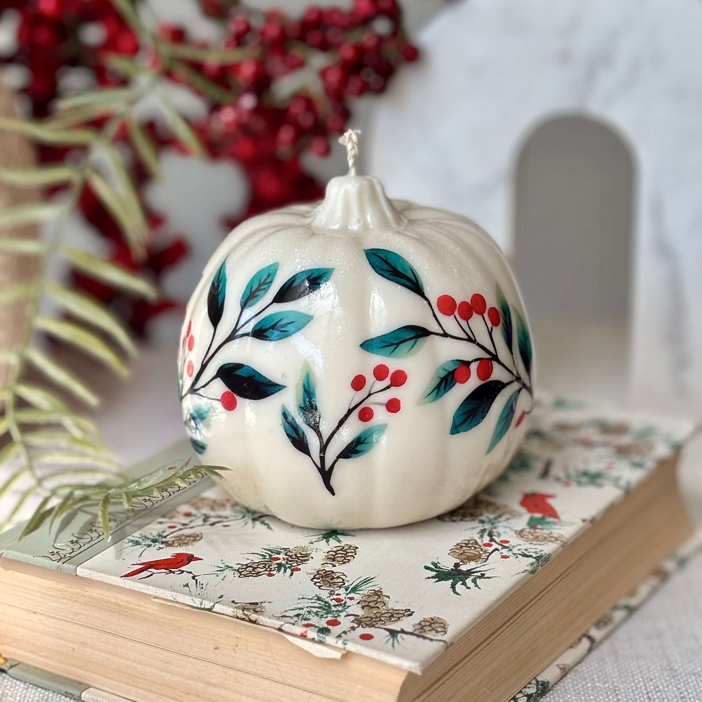 Handmade pumpkin candle with painted floral and berry design placed on a holiday-themed book, ideal for Thanksgiving, Halloween, cozy fall birthdays or Secret Santa gifts