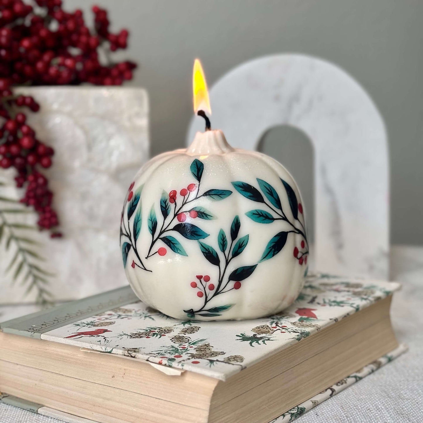 Soy pumpkin candle with painted floral design, burning on a decorated book and surrounded by red berries and holiday decor — ideal for Christmas, Thanksgiving or winter birthdays