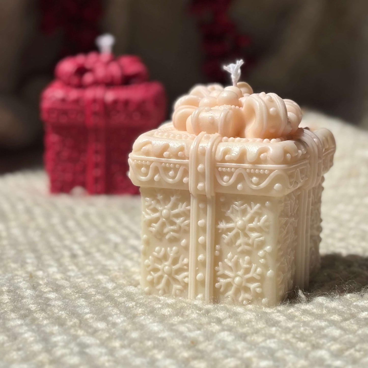 Beige holiday candle shaped like a gift box with red festive candle in background.