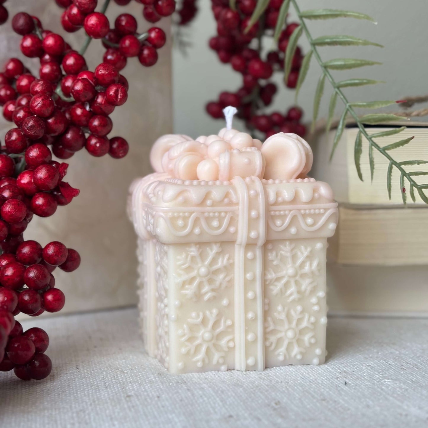 Handmade beige Christmas candle shaped like a gift box surrounded by red berries and greenery.