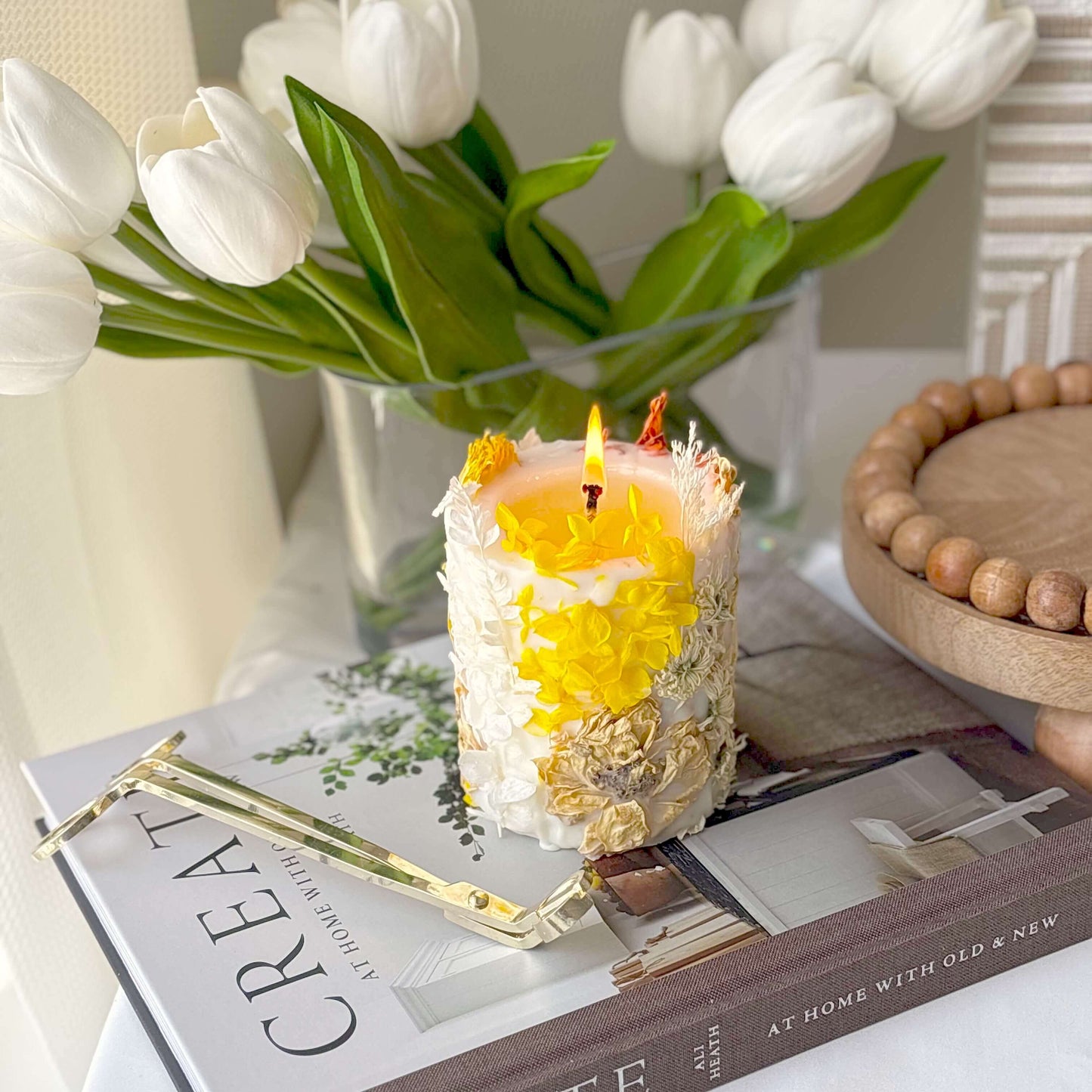 Burning handmade soy candle with yellow and white dried flowers, styled on books and tulips. Botanical decor piece made in California.
