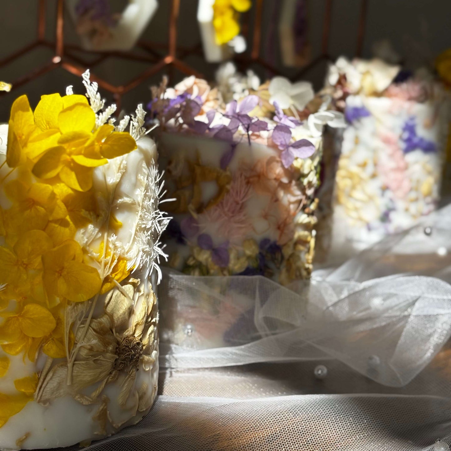 Close-up of handmade botanical soy candles with pressed yellow, purple, and white flowers, illuminated by warm sunlight — elegant artisan decor and gift