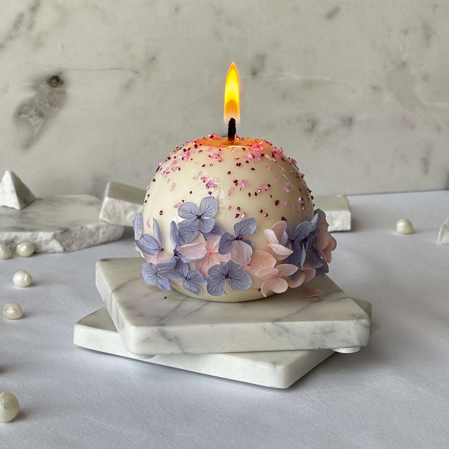Lit round flower candle with delicate pink sparkles and pressed petals on white marble base