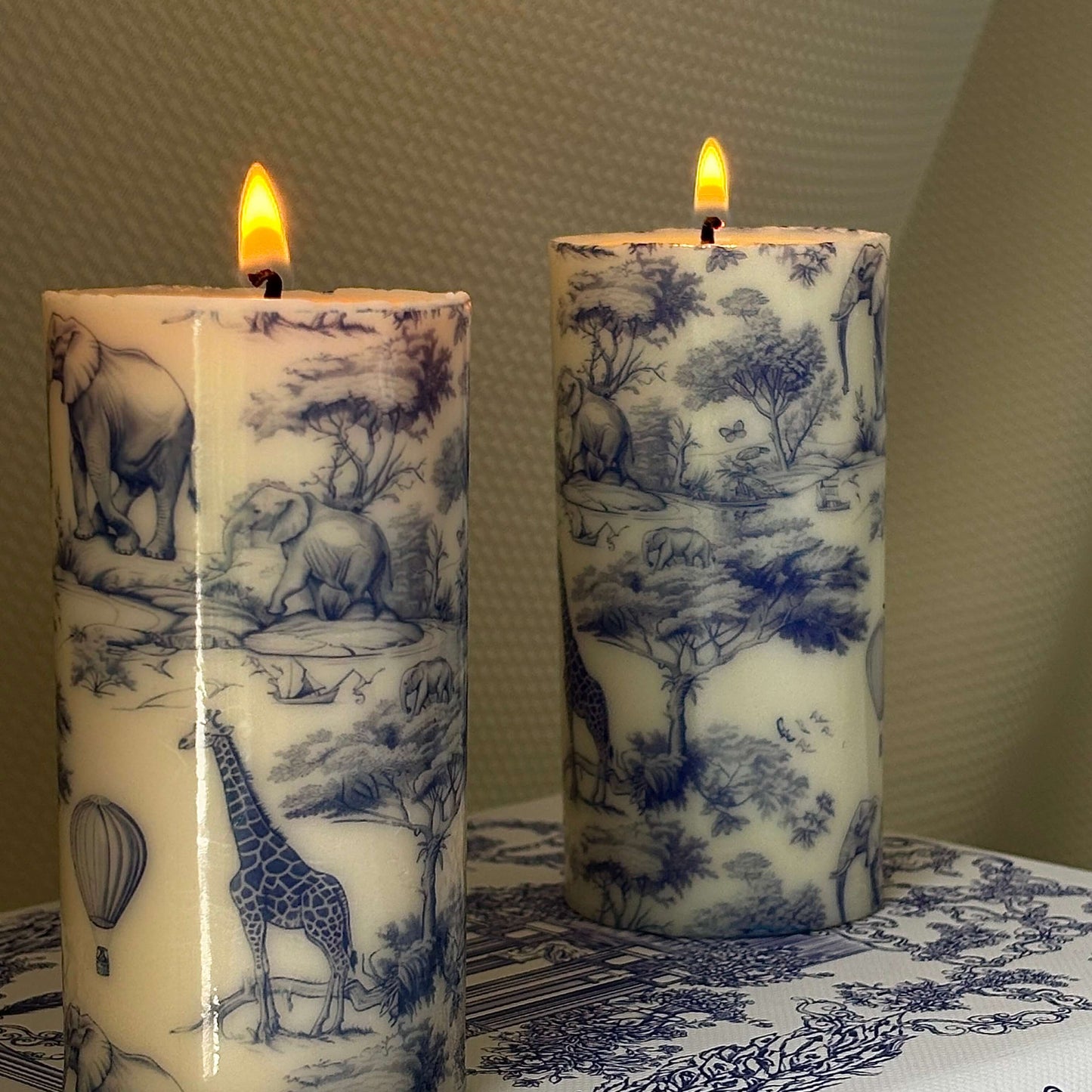 Close-up of lit handmade candles with glossy finish and detailed Toile de Jouy safari print featuring giraffes and elephants.