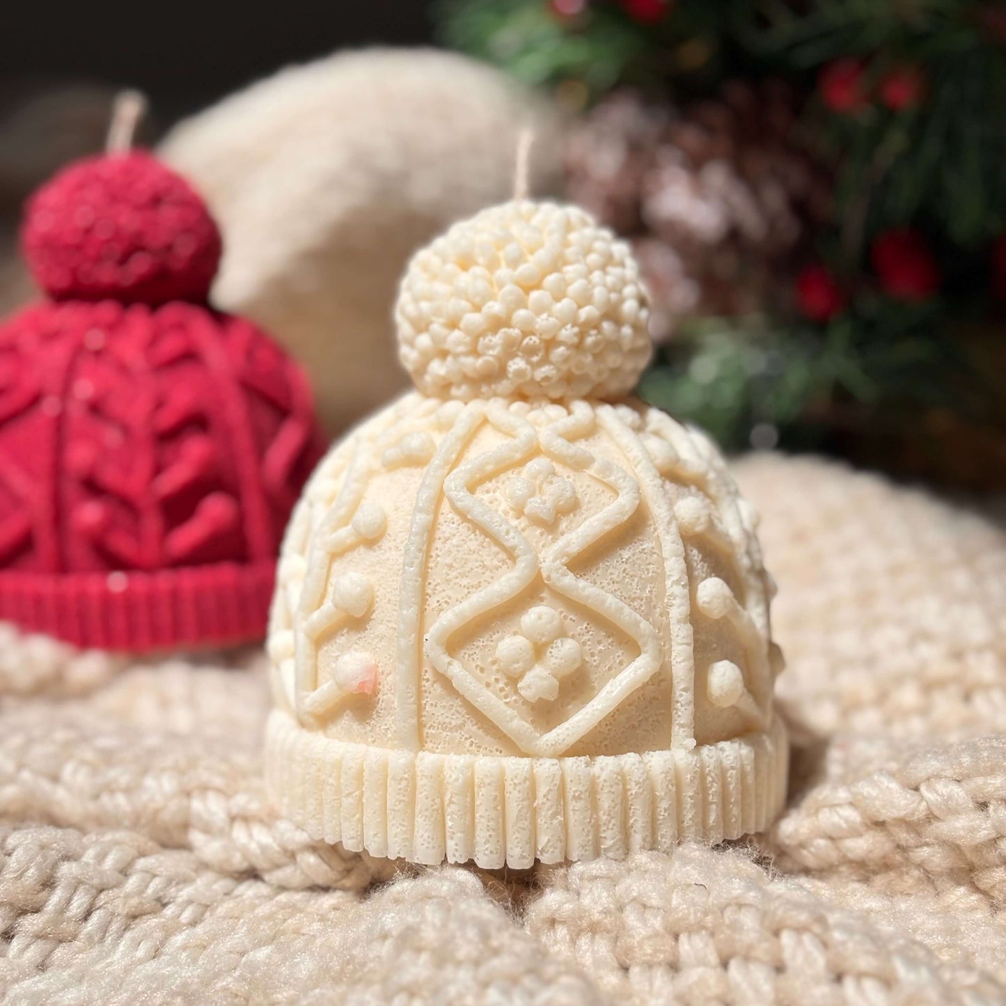 Cream knit hat candle on soft knitted fabric with blurred red version in the background, styled for cozy winter gift or Secret Santa present