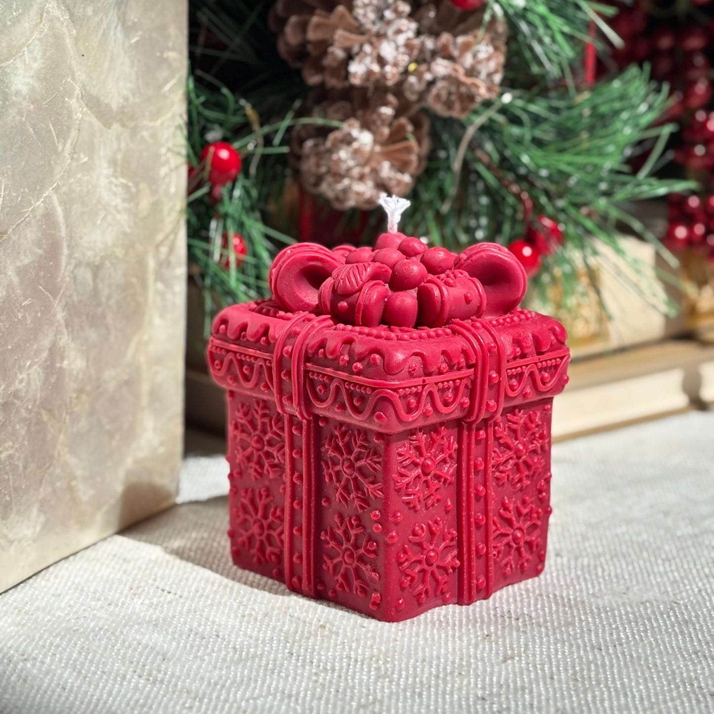 Decorative red gift box candle with holiday pine and Christmas tree in background.