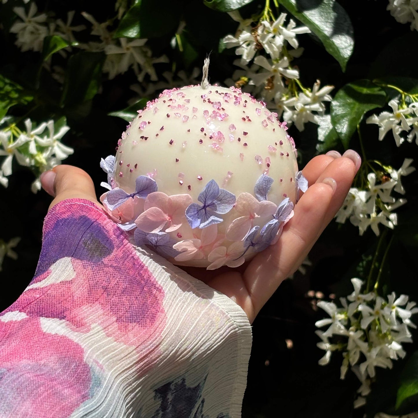 Hand holding round floral candle with pink sprinkles and purple hydrangea flowers, garden background