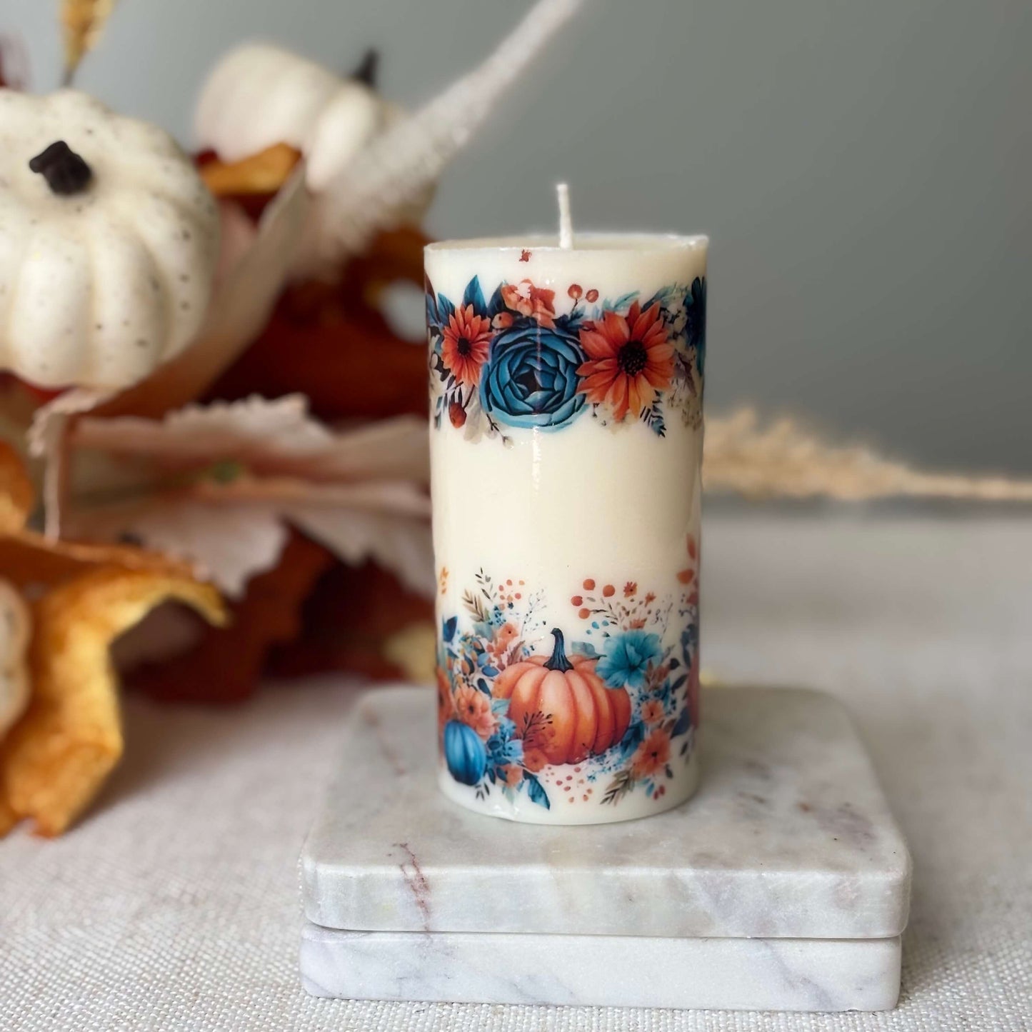 Glossy floral pumpkin candle on marble base surrounded by autumn leaves and white pumpkins
