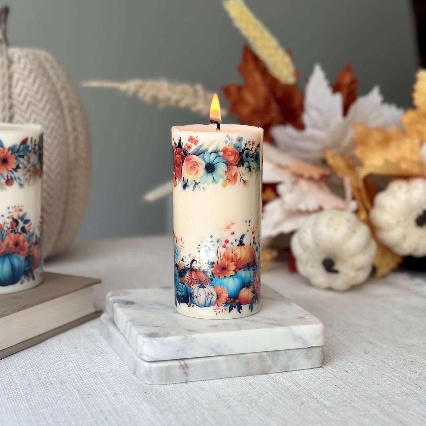 Handmade glossy pumpkin candle with autumn floral design on marble base surrounded by seasonal decor