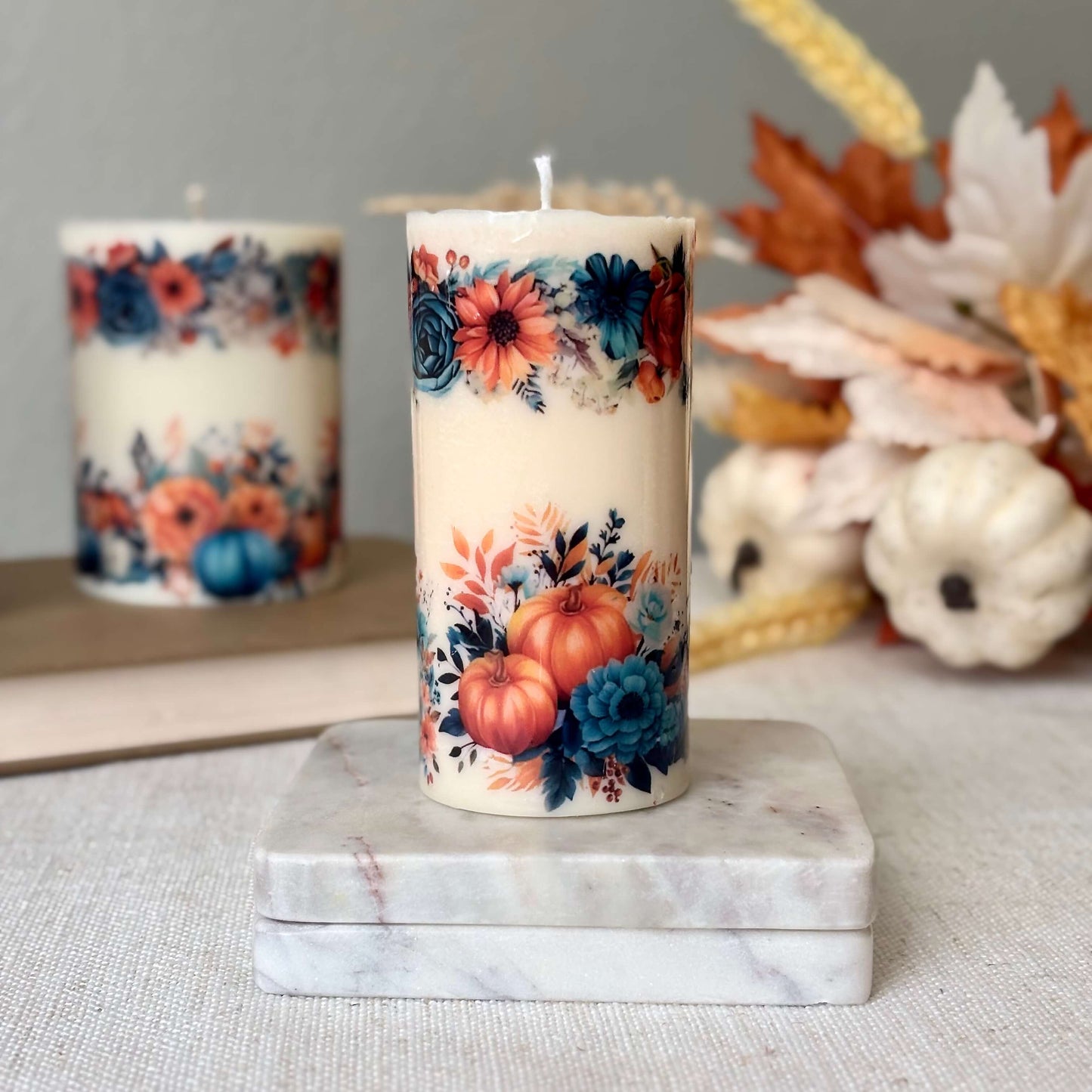Glossy pumpkin candle with floral autumn design on marble base with another candle in background