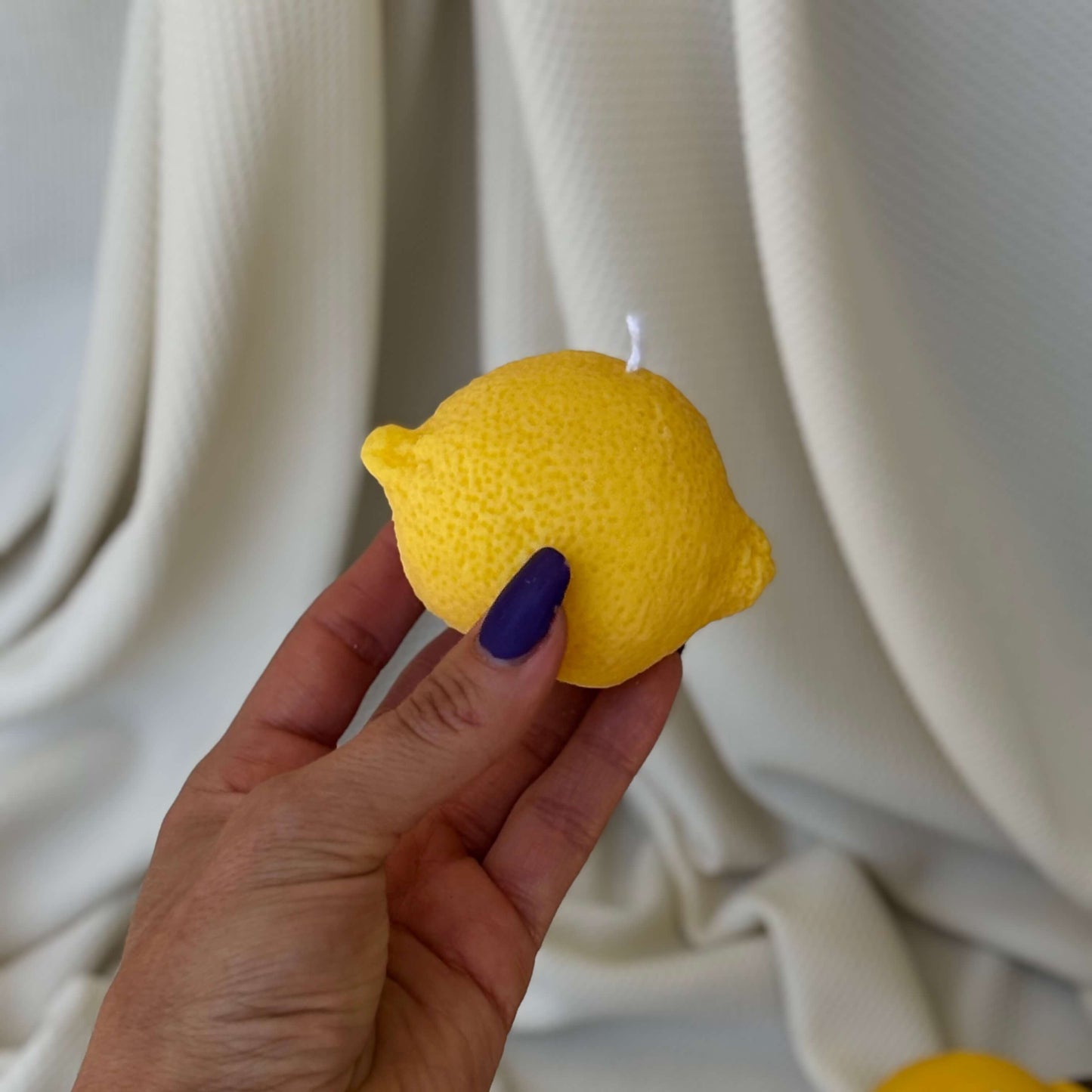 Hand holding a horizontal lemon-shaped candle in front of soft white background. Realistic soy wax decor.
