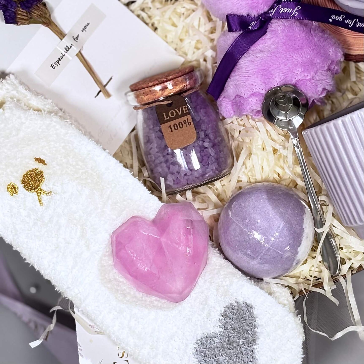Flatlay of lavender spa gift items including heart soap, bath bomb, plush socks, bath salt, teddy candle, spoon and ceramic mug on shredded filler