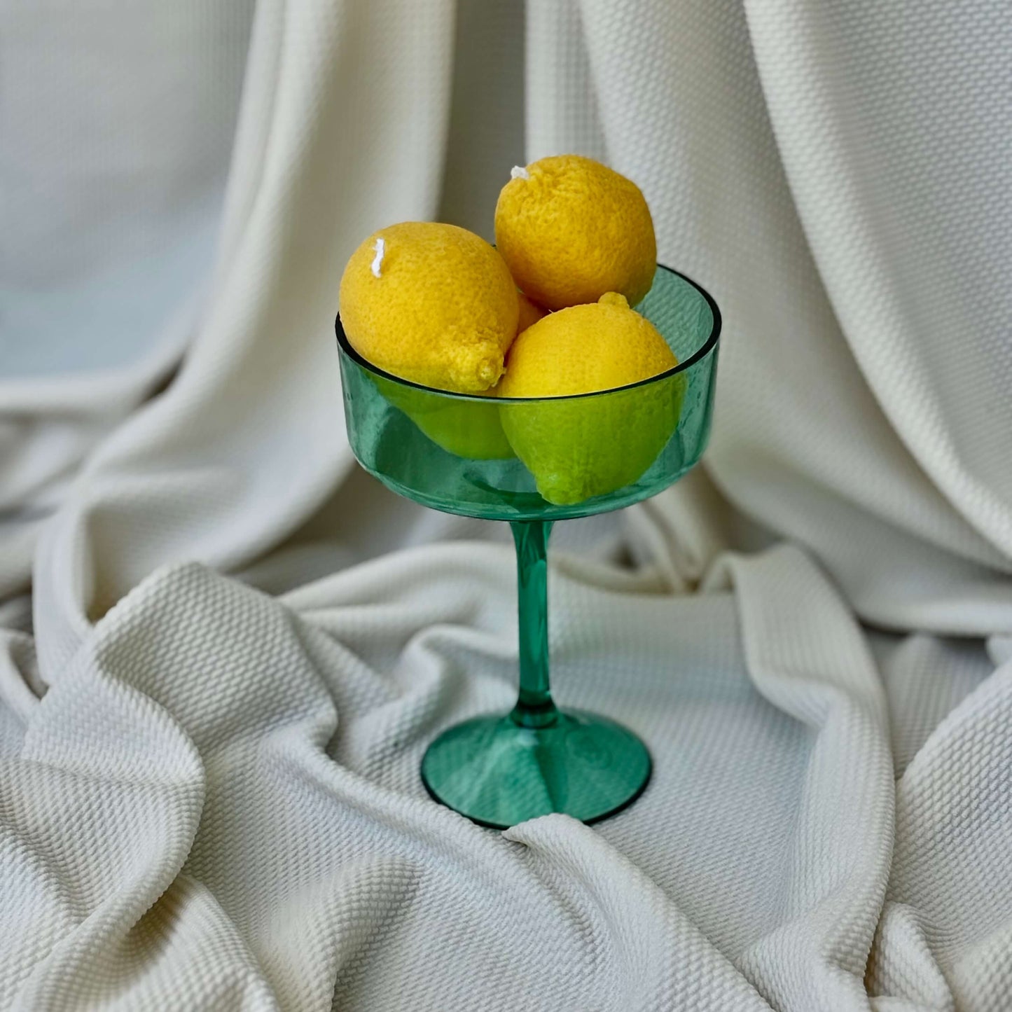 Lemon candles stacked in green glass bowl on white fabric. Decorative citrus-shaped candles for aesthetic setup.