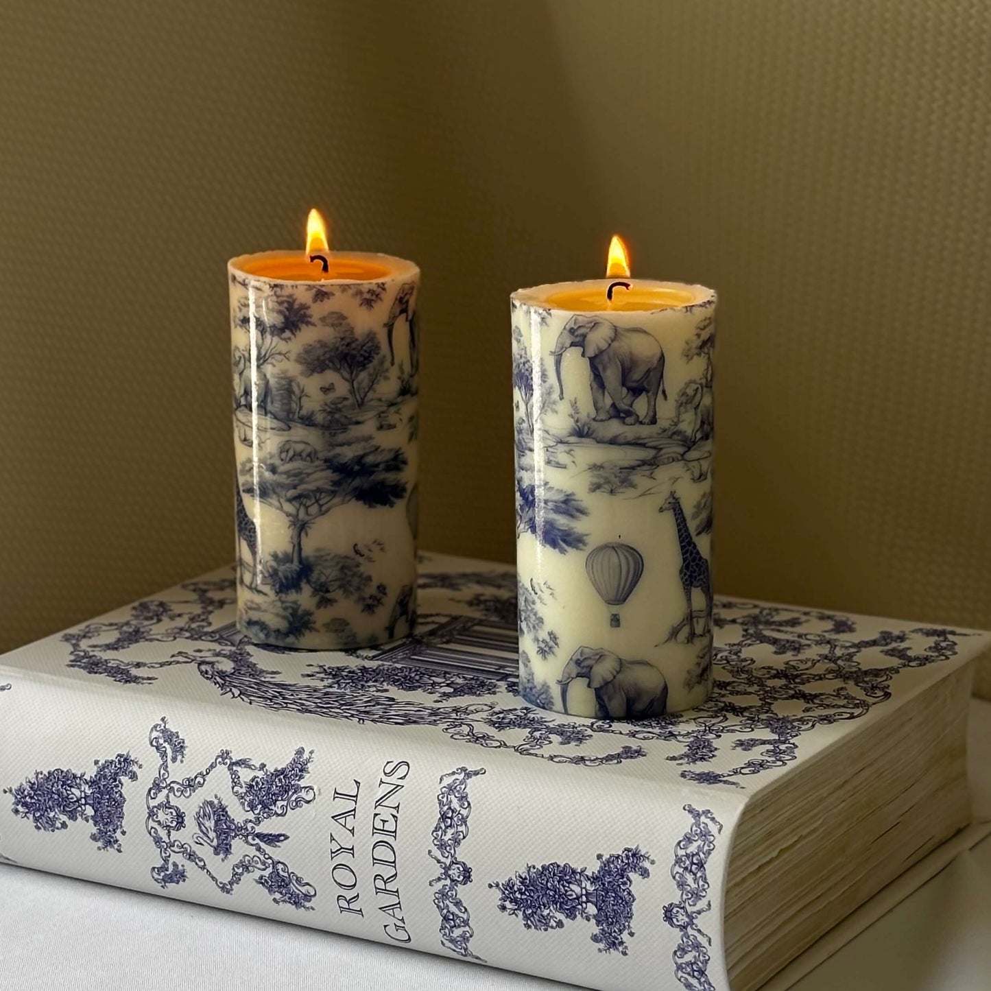Set of two lit glossy pillar candles with Toile de Jouy print, styled on decorative book, perfect for Afro-inspired coffee table décor.