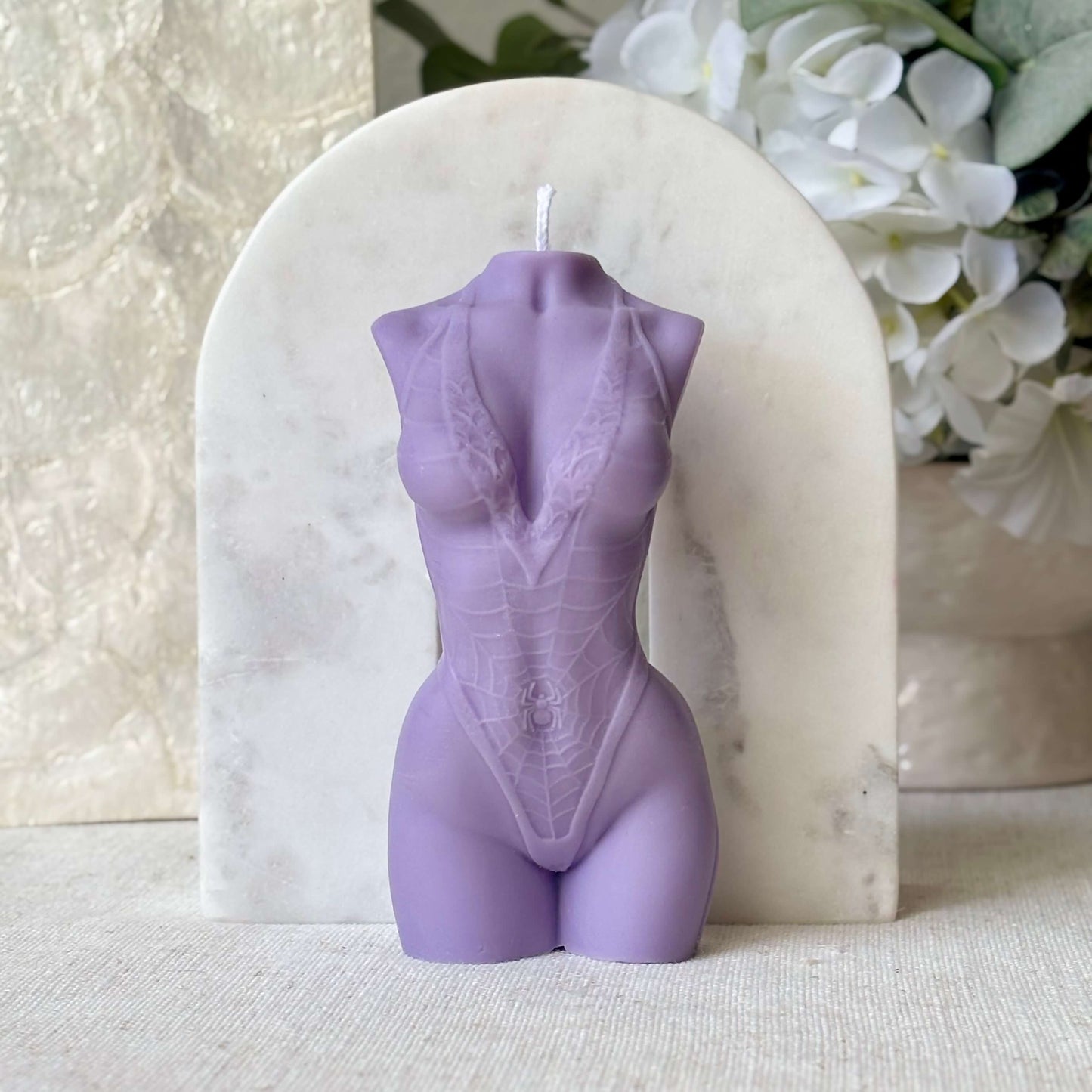 Purple female torso candle handmade from soy wax, artistic body sculpture accent with white flowers in background.