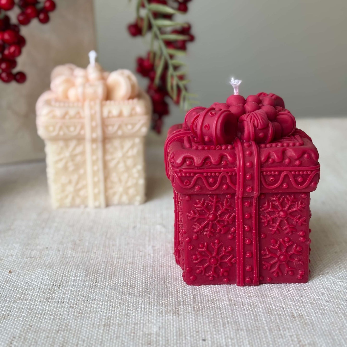 Red soy candle shaped like a festive gift box with beige candle in background and holiday greenery.
