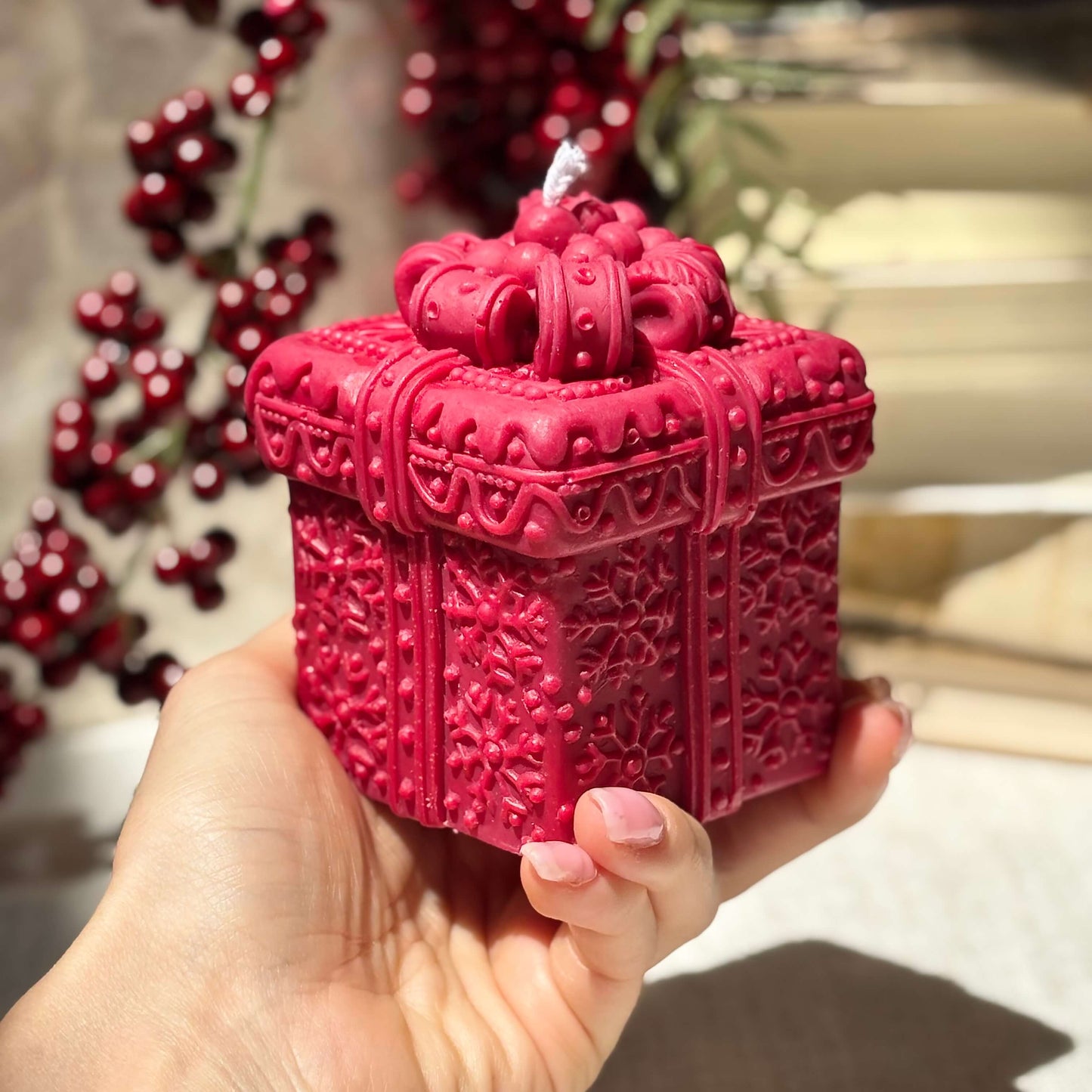 Red handmade candle shaped like a gift box, held in hand with red berries in background.