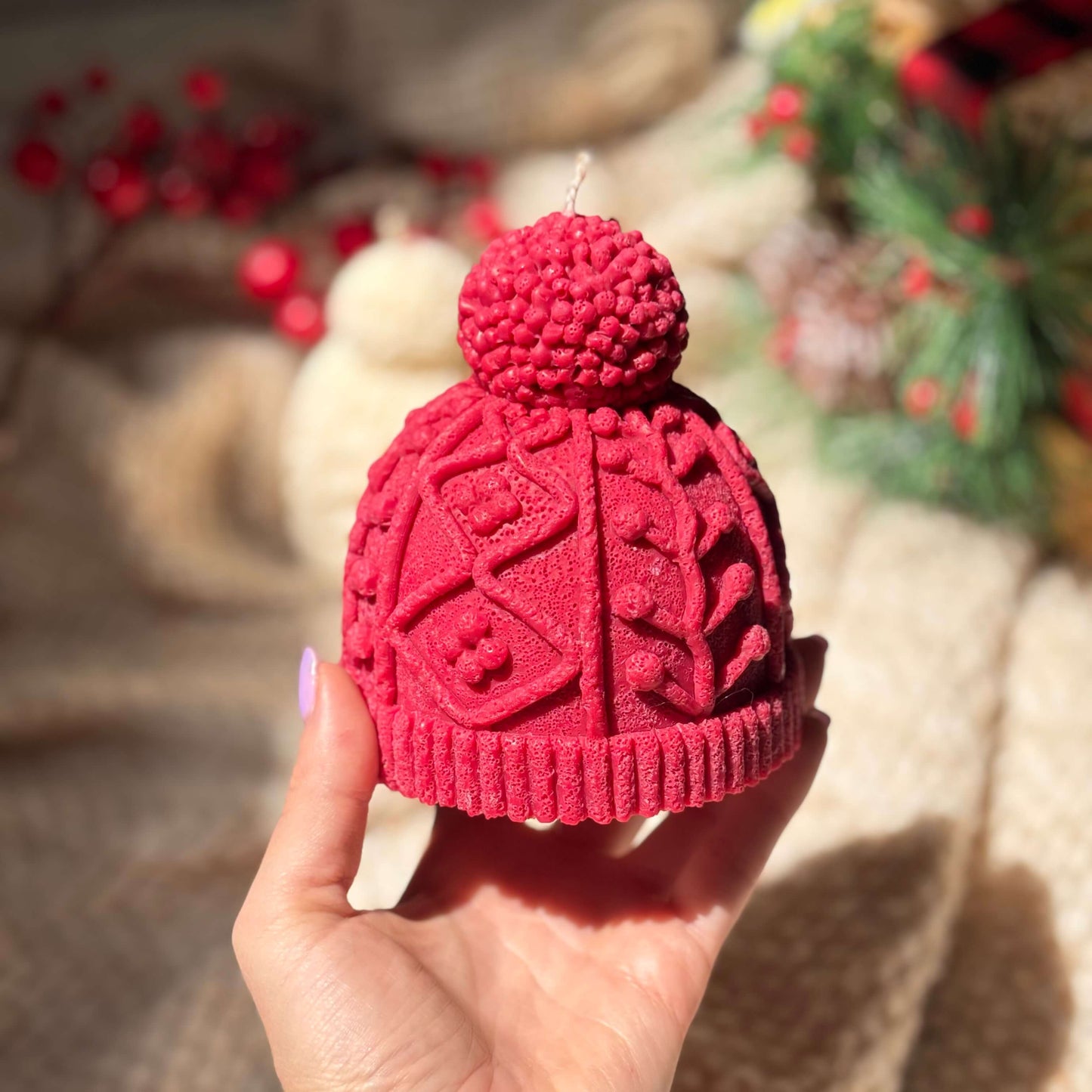 Hand holding a red handmade candle shaped like a cozy knit hat with blurred Christmas greenery and soft light in the background