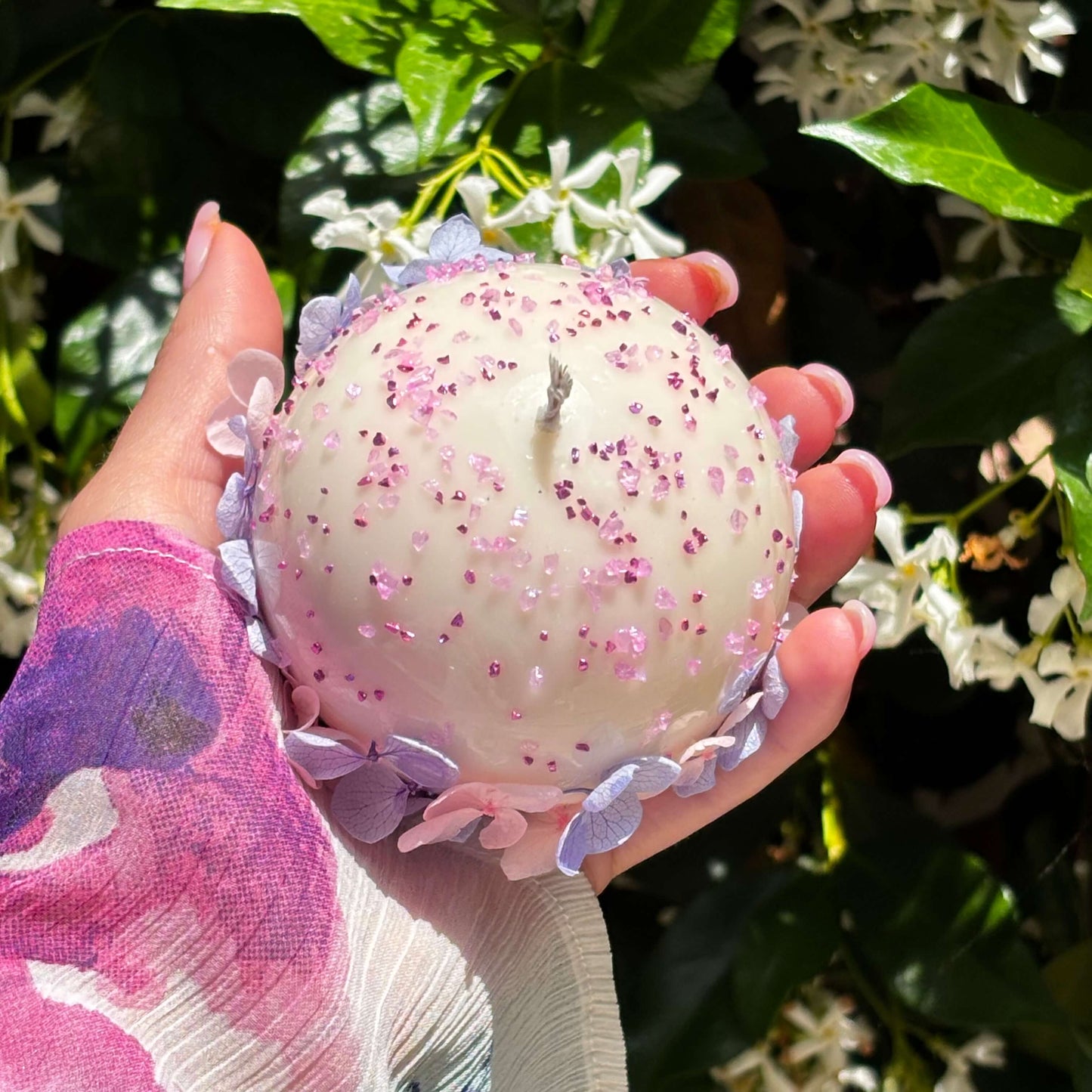 Top view of round floral candle with pink glitter and hydrangea flowers held in hand, garden background