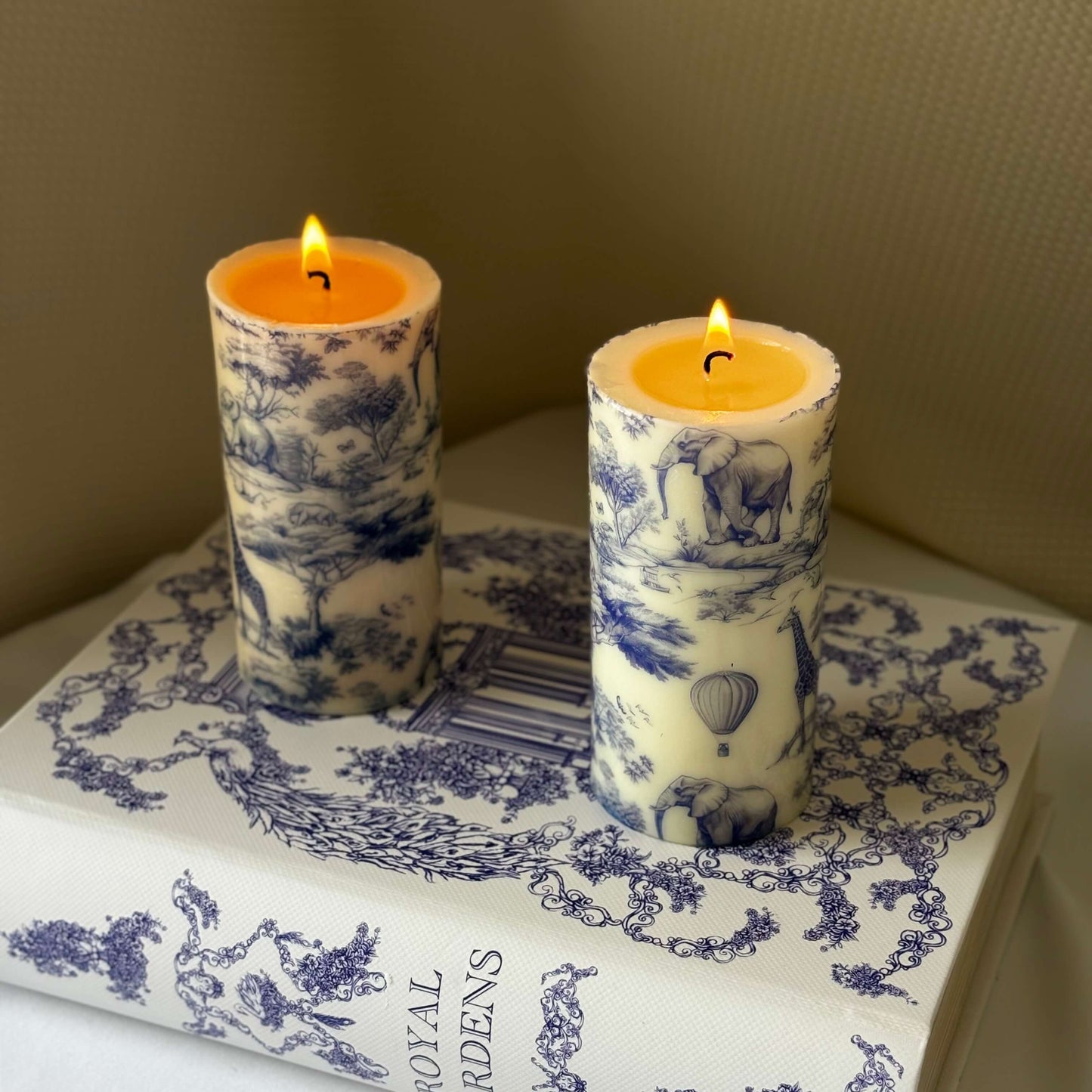 Two lit glossy candles with Toile de Jouy safari print placed on Royal Gardens decor book, perfect for elegant Afro-style table setting.