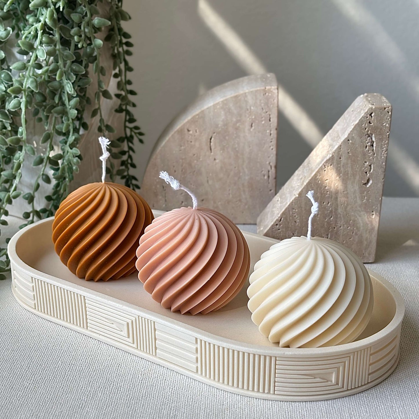 Three sculptural swirl candles in peach, ivory and honey colors displayed on a decorative tray with greenery background.