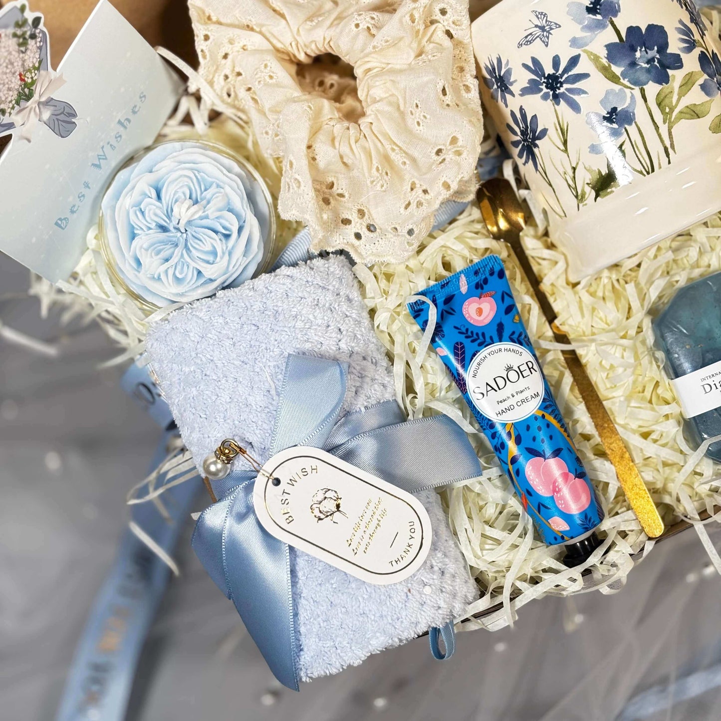 A beautifully arranged blue-themed spa gift box featuring a floral ceramic mug, hand cream, soft towel with ribbon, golden spoon, handmade soap, scrunchie, and decorative flower, nestled in shredded filler.