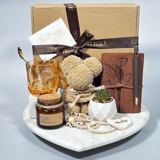 Gift box with a towel folded into a teddy bear shape, amber glass cup with golden spoon, soy candle in a jar, mini succulent in a white ceramic pot, brown notebook with pen, and hair ties, all arranged on a white marble tray.