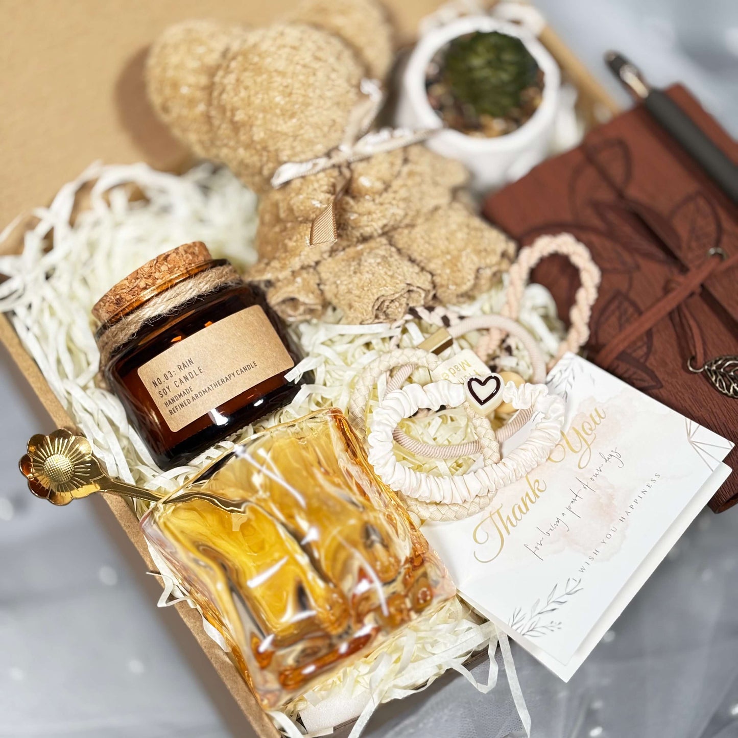 Top view of a gift box featuring a soy candle in an amber jar, sculpted glass cup with gold spoon, towel folded into a teddy bear shape, set of hair ties, thank-you card, brown notebook with pen, and a mini succulent in a white pot.