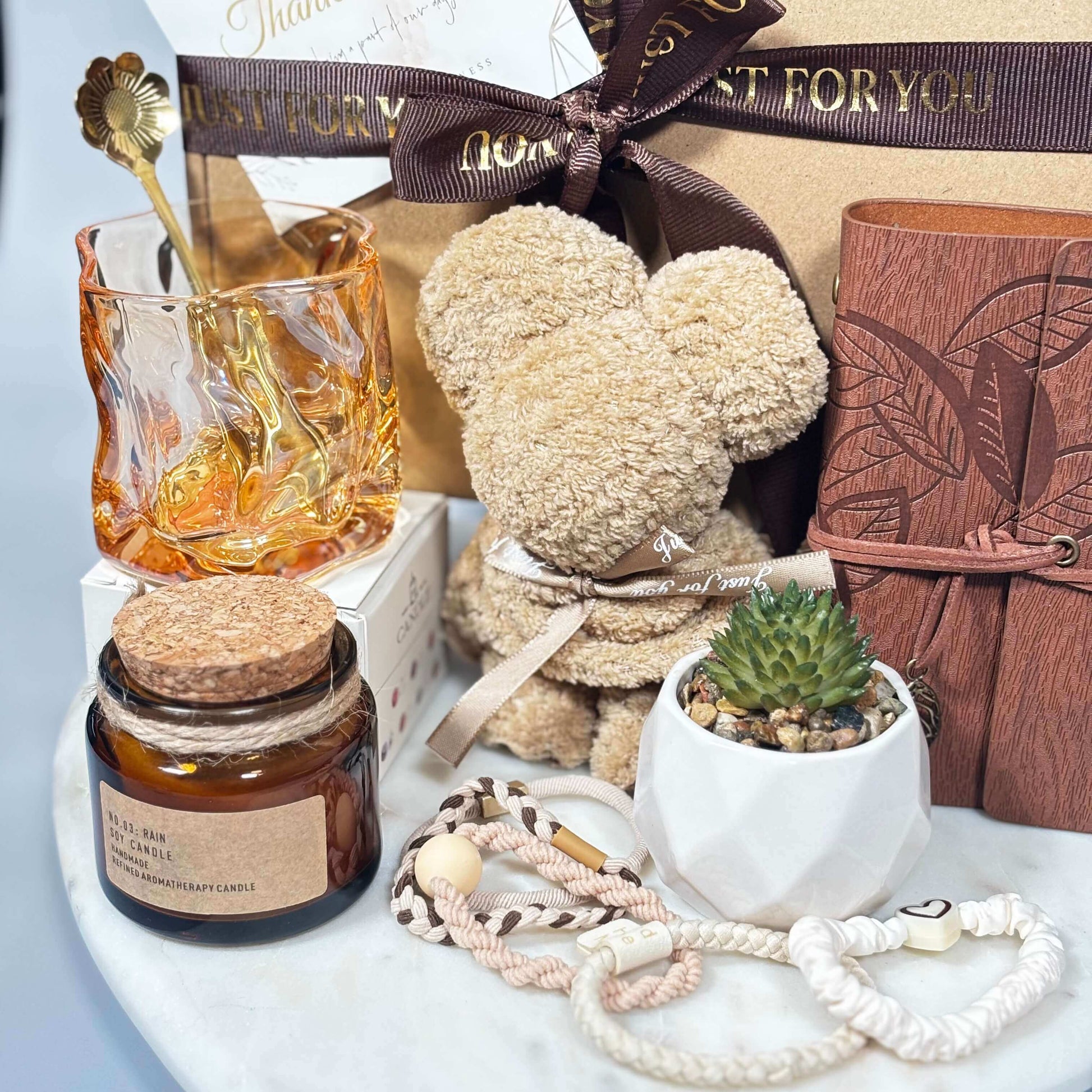 Close-up of a cozy gift box featuring a towel folded into a teddy bear shape, soy candle in amber jar, amber glass with gold spoon, mini succulent in a ceramic pot, leather-style notebook with pen, and a set of handmade hair ties.