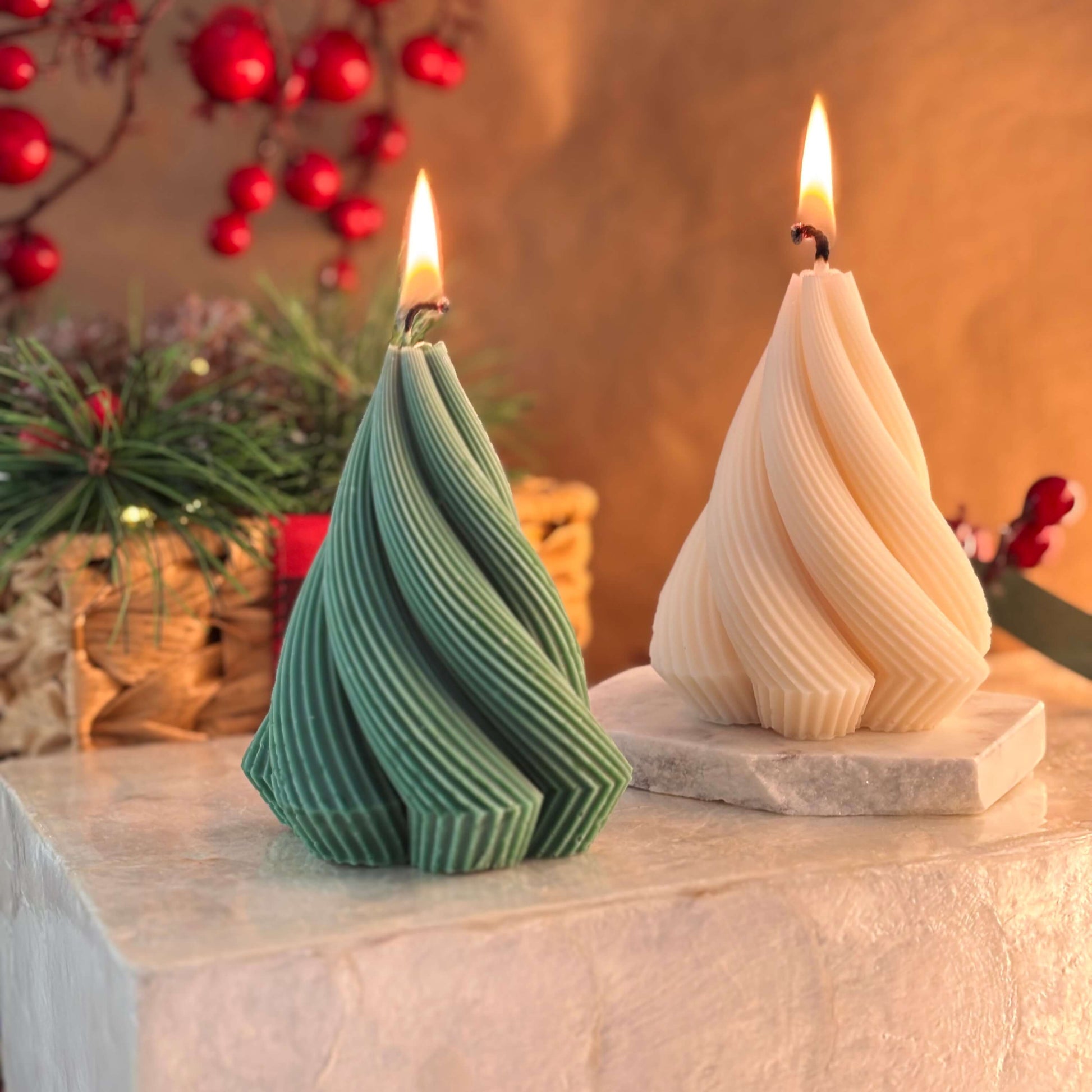 Green sculpted soy wax candle in the shape of a Christmas tree with elegant twisted design, next to an ivory version on a festive stone block

