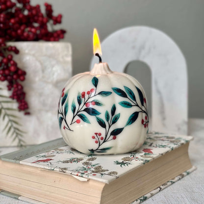 Soy pumpkin candle with painted floral design, burning on a decorated book and surrounded by red berries and holiday decor — ideal for Christmas, Thanksgiving or winter birthdays


