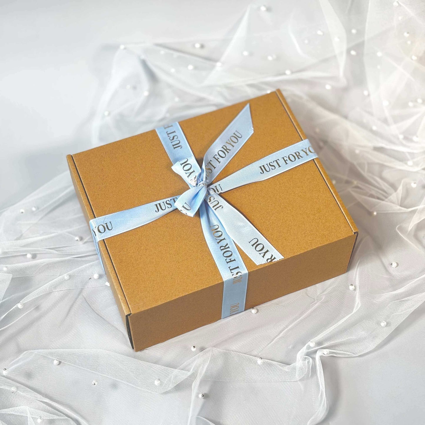Closed kraft gift box tied with a light blue “Just For You” ribbon, placed on delicate white tulle with pearl details.
