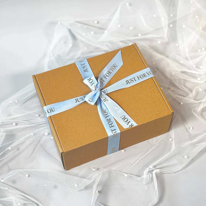 Closed kraft gift box tied with a light blue “Just For You” ribbon, placed on delicate white tulle with pearl details.