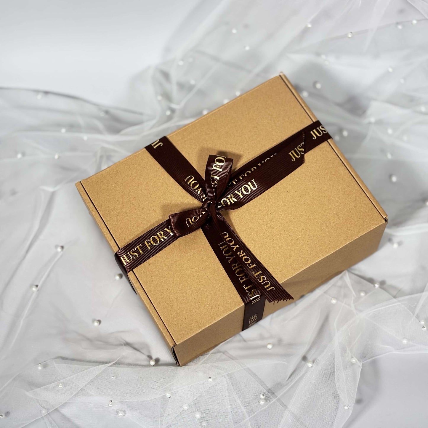 Closed kraft gift box tied with a brown “Just For You” ribbon, placed on soft white tulle with pearl accents.
