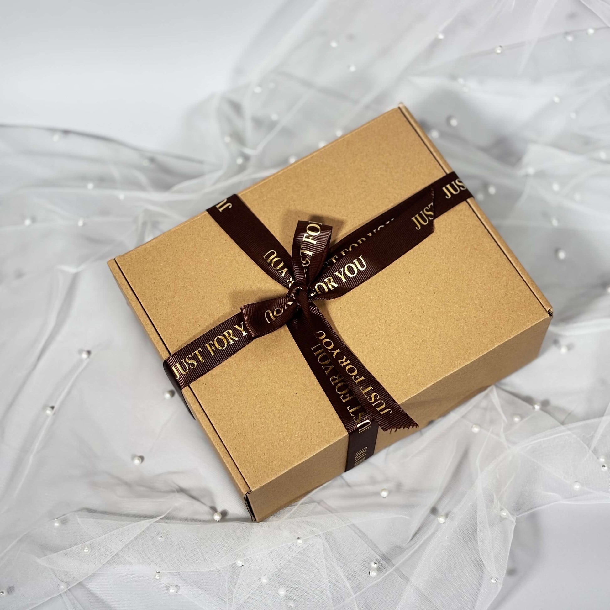 Closed kraft gift box tied with a brown “Just For You” ribbon, placed on soft white tulle with pearl accents.
