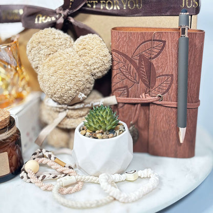 Close-up of a gift box featuring a towel folded into a teddy bear, mini succulent in a white ceramic pot, brown embossed notebook with pen, soy candle, and a set of handmade hair ties arranged on a white marble surface.