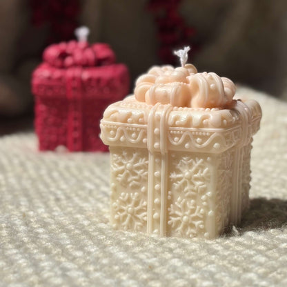 Beige holiday candle shaped like a gift box with red festive candle in background.