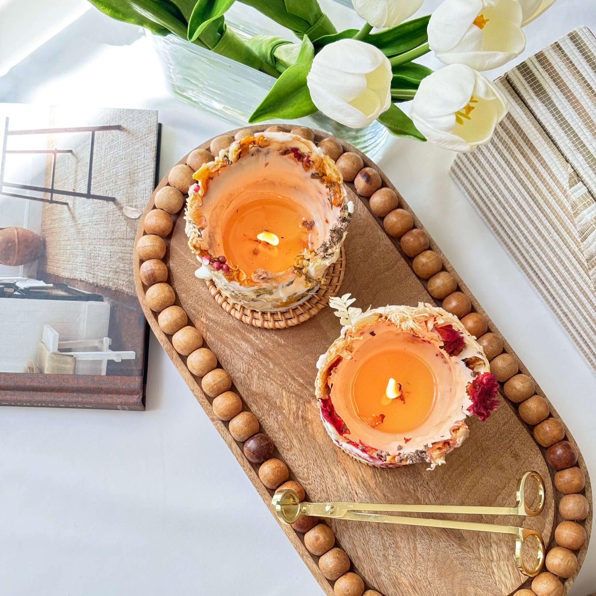 Two handmade botanical soy candles with dried flowers, burning on a wooden tray with candle tools and white tulips in the background.