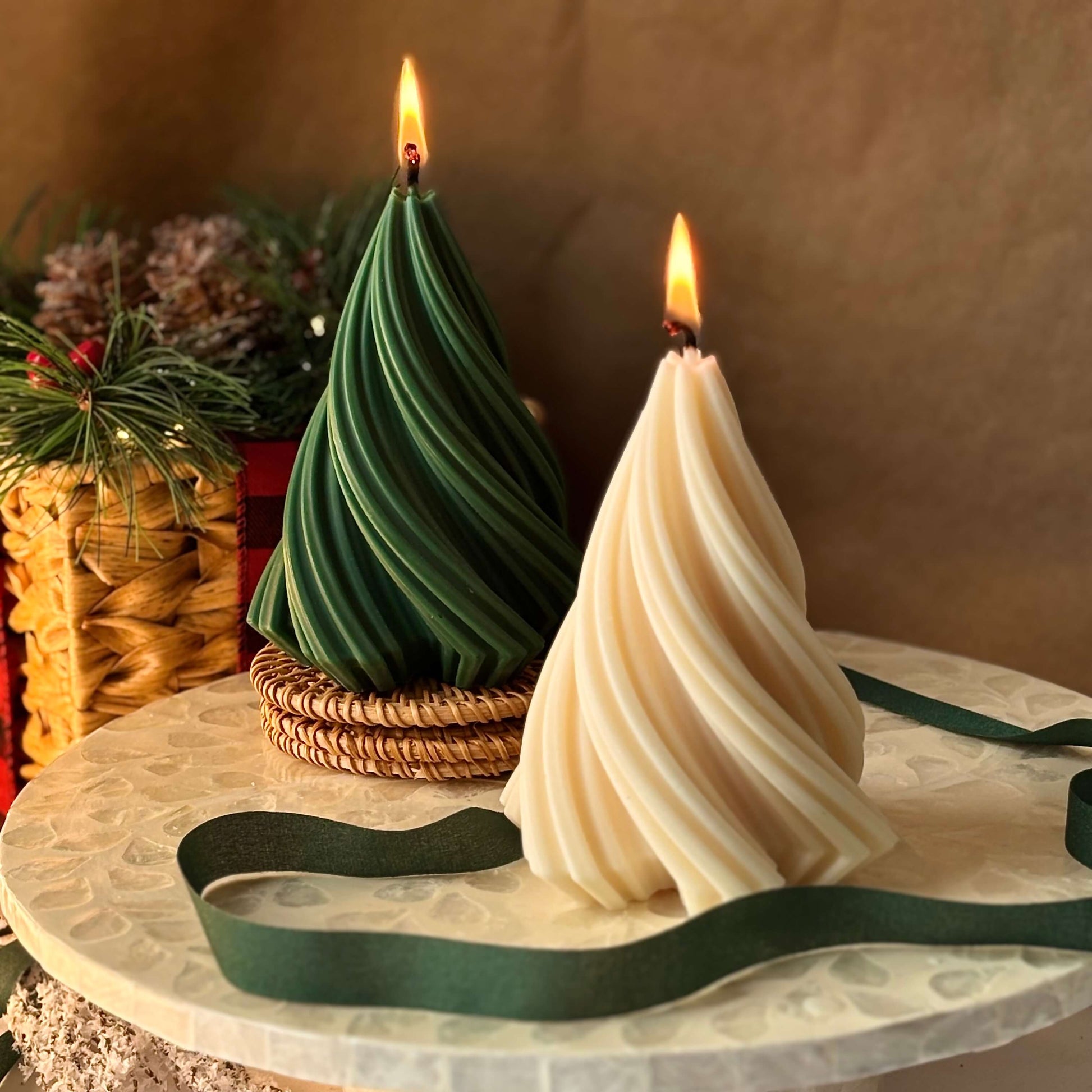 Spiral Christmas tree candles in green and cream, lit on a marble surface with ribbon and holiday greenery in the background