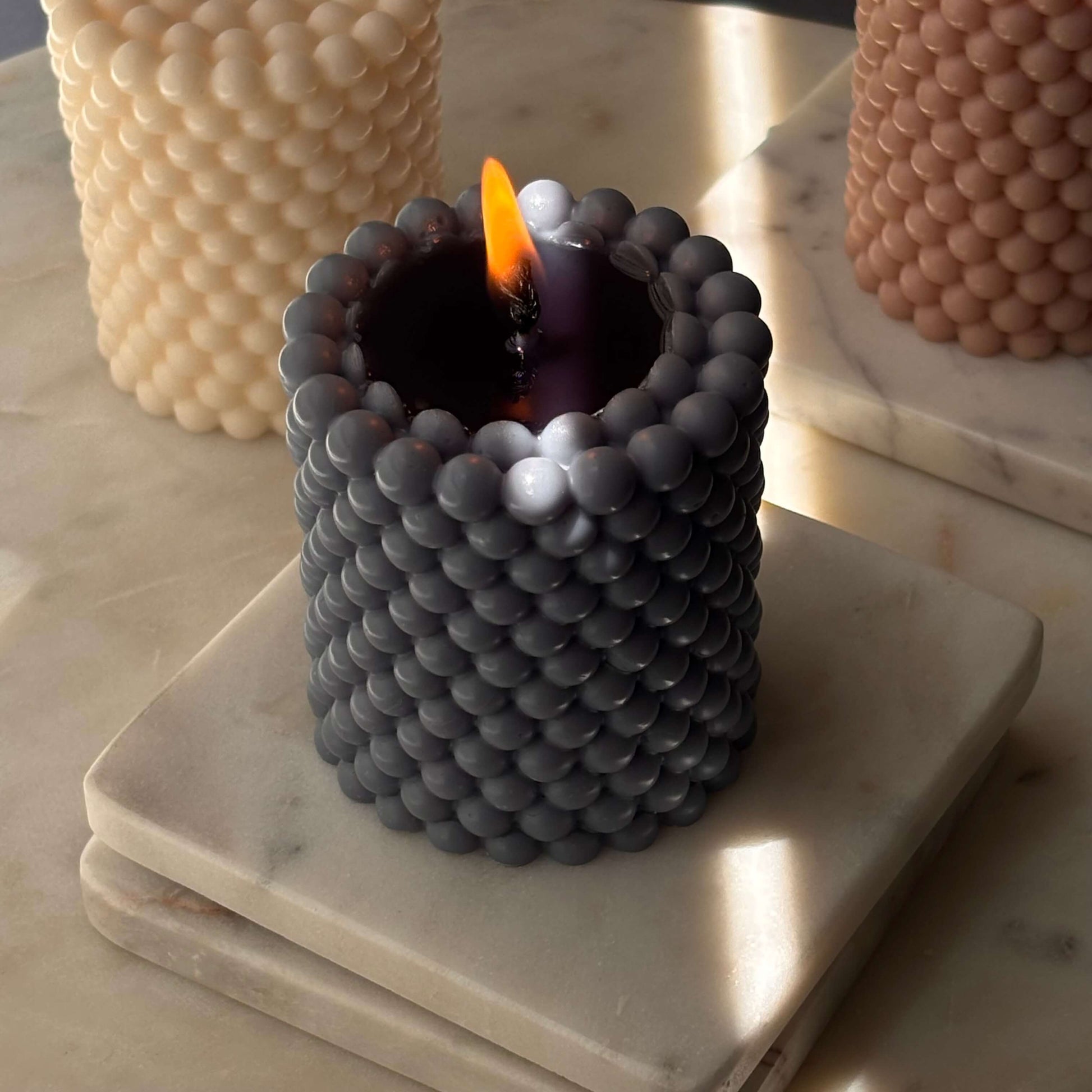 Burning charcoal-toned candle with pearl texture — soy wax sculpture with moody glow on marble.

