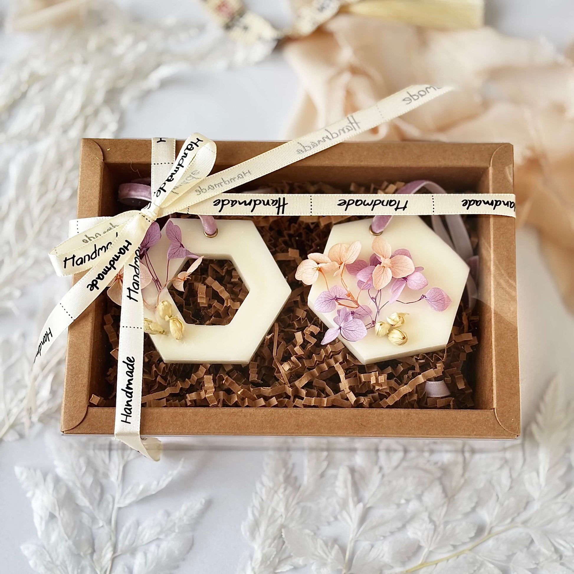 Two handmade soy wax sachets decorated with pink and lavender dried flowers, arranged in a kraft gift box with paper filler and “Handmade” ribbon.