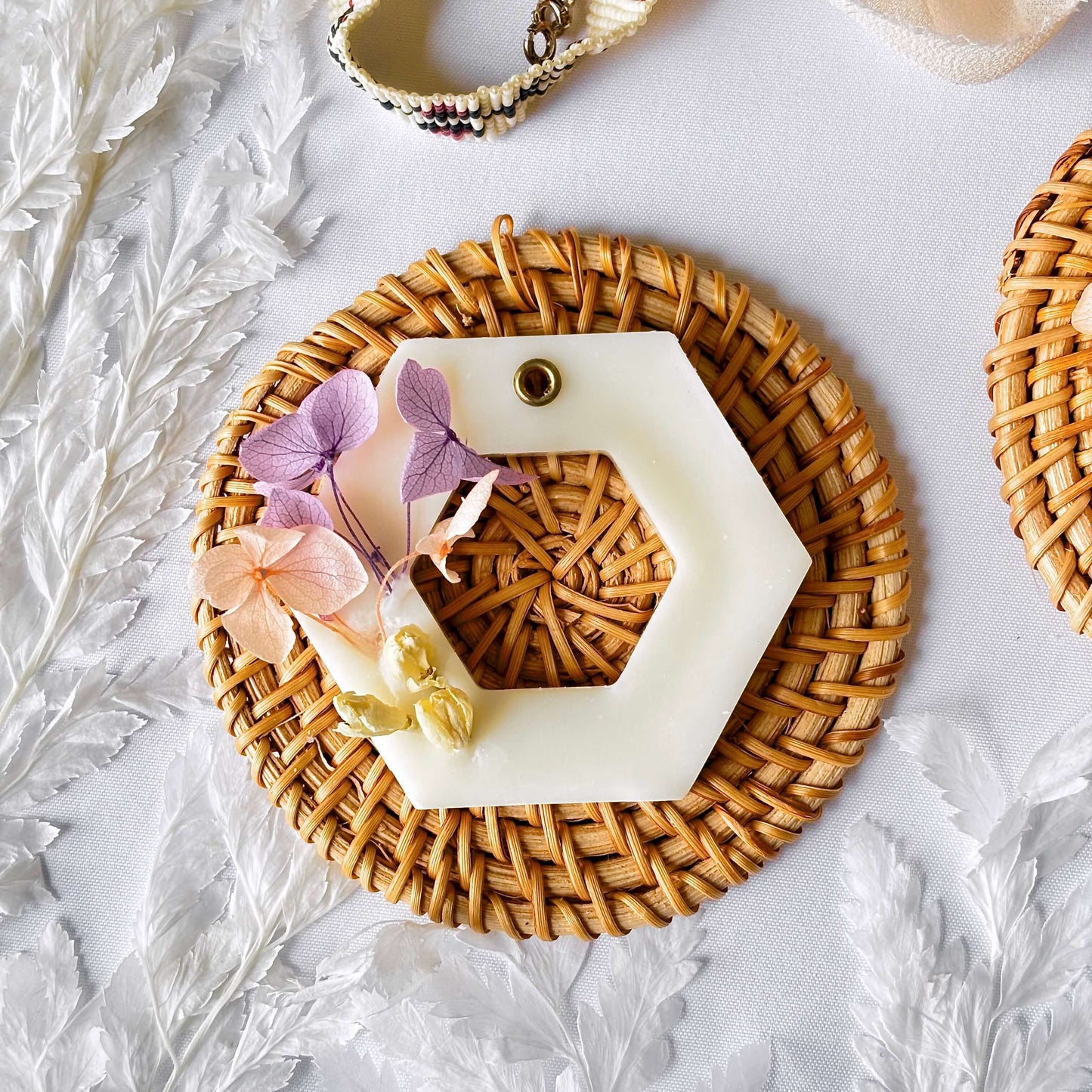 Hexagon-shaped soy wax sachet with dried pink and purple flowers and twine loop, styled flat on a woven rattan base with white botanical accents.