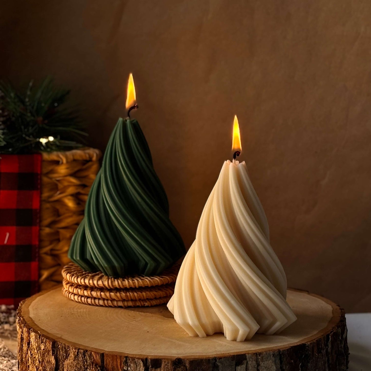 Two lit Christmas tree-shaped candles, green and beige, placed on a wooden slice with warm festive decor in the background