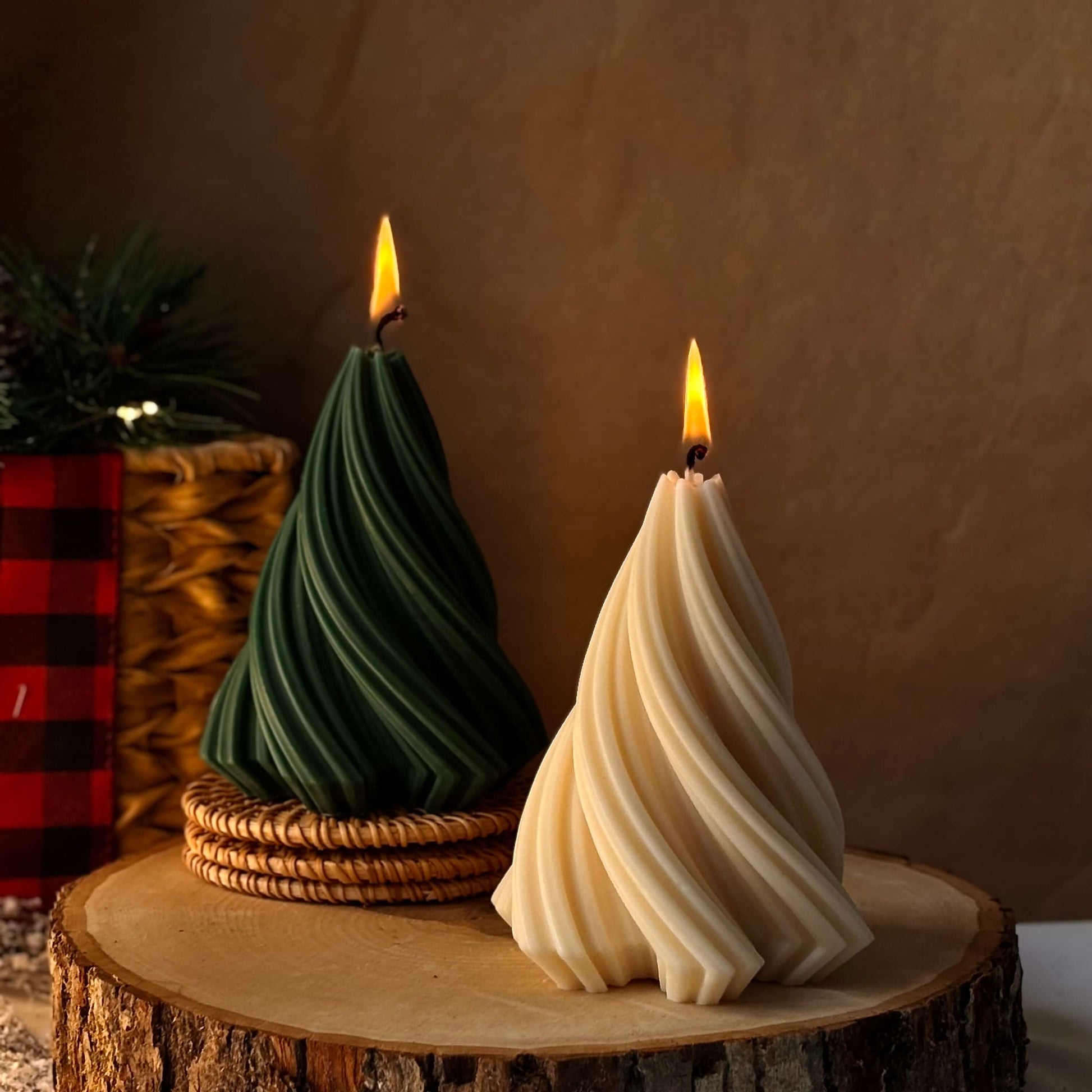 Two lit Christmas tree-shaped candles, green and beige, placed on a wooden slice with warm festive decor in the background