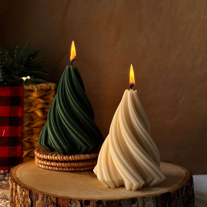 Two lit Christmas tree-shaped candles, green and beige, placed on a wooden slice with warm festive decor in the background