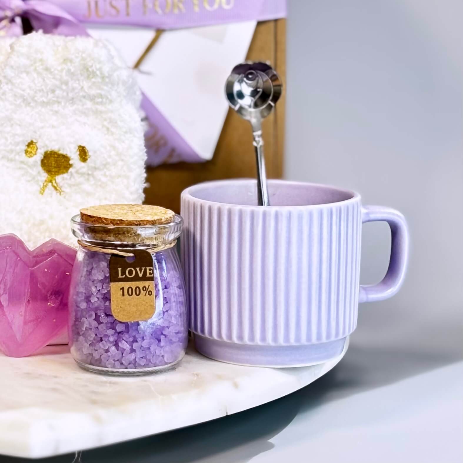 Close-up of lavender ceramic mug with stainless steel flower spoon and jar of purple bath salt on marble tray
