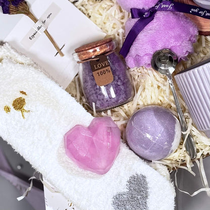 Flatlay of lavender spa gift items including heart soap, bath bomb, plush socks, bath salt, teddy candle, spoon and ceramic mug on shredded filler

