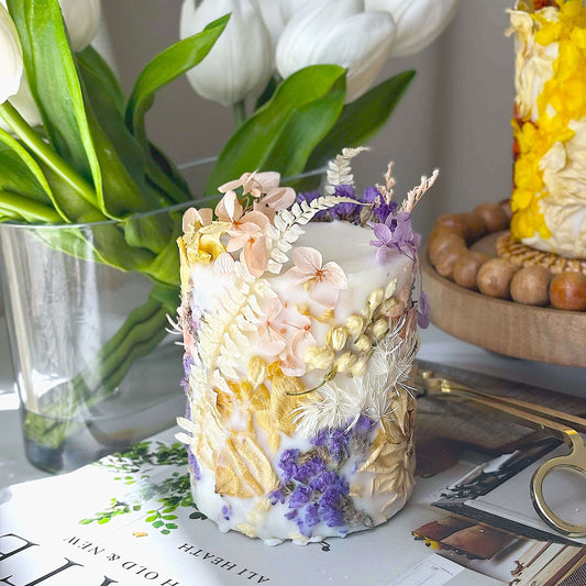 Handmade soy wax candle with pressed pastel flowers in pink, purple, and cream tones. Botanical decorative piece styled on a table with tulips and gold scissors.