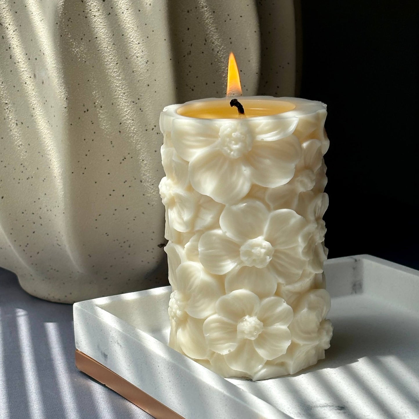 Lit floral soy pillar candle in natural light beside textured ceramic vase	