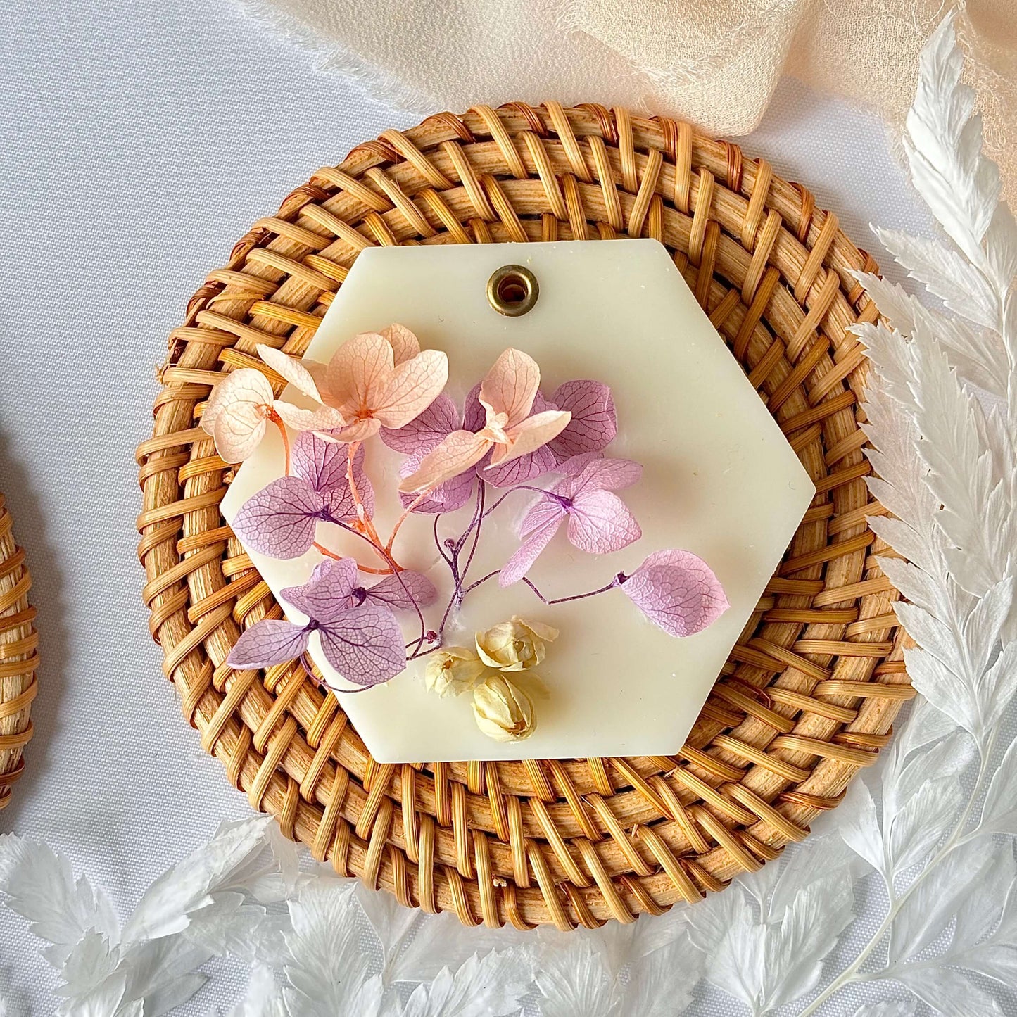 Single handmade soy wax sachet decorated with pink and lavender dried flowers, styled on a round rattan mat with white botanical accents in the background.